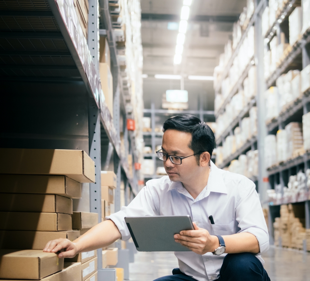A man in glasses references a tablet while checking items on a warehouse floor.