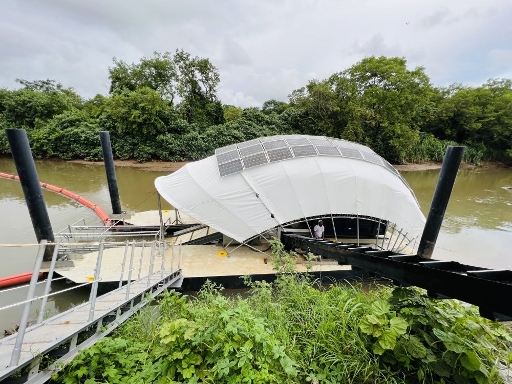 A mechanical wheel topped with a tarp-like cover tiled in solar panels sits in a green river surrounded by green foliage