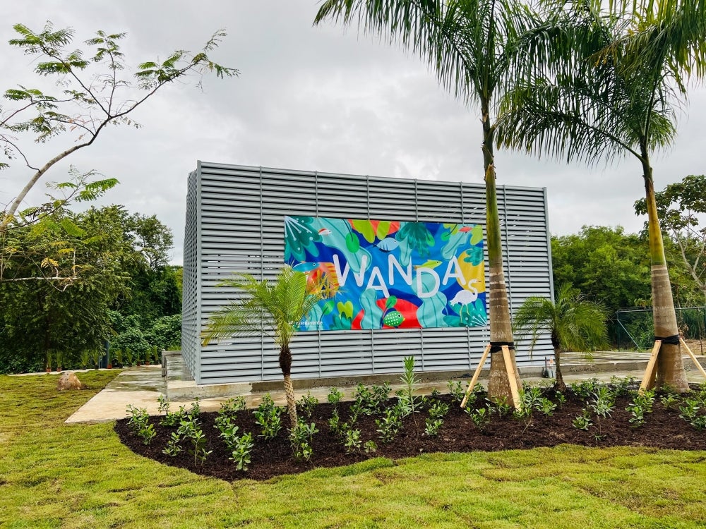 A steel structure sits behind palm trees and is adorned with a bright, multicolored banner reading 