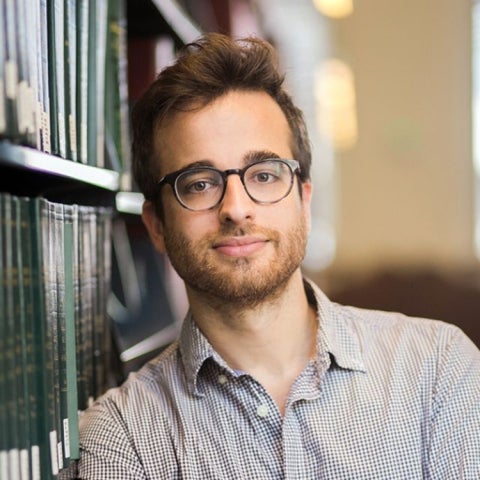 Michael Goard wears a collared shirt and leans against a bookshelf