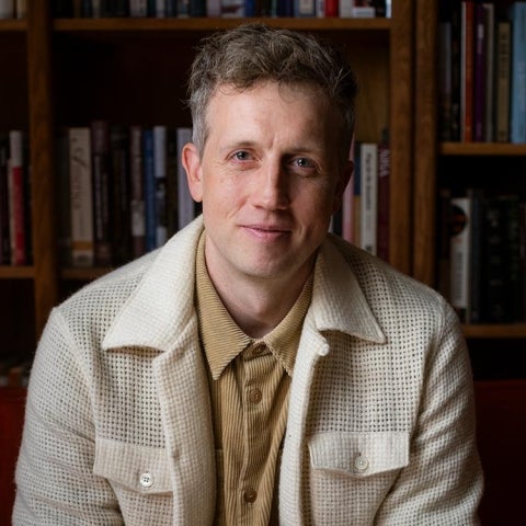 Joseph Blankholm wears a collared shirt and a jacket while sitting in front of a bookshelf