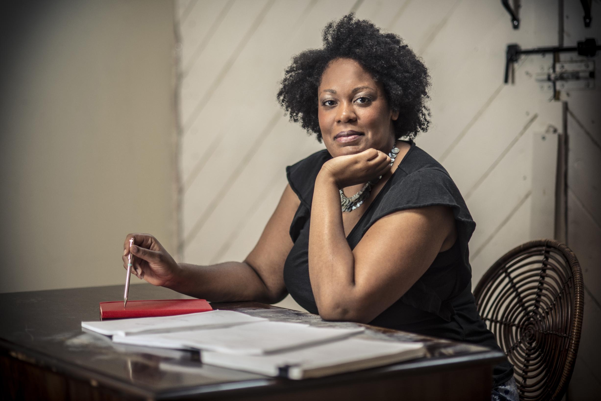 Playwright Jacqueline Lawton sits at a desk holding a pen above a red notebook