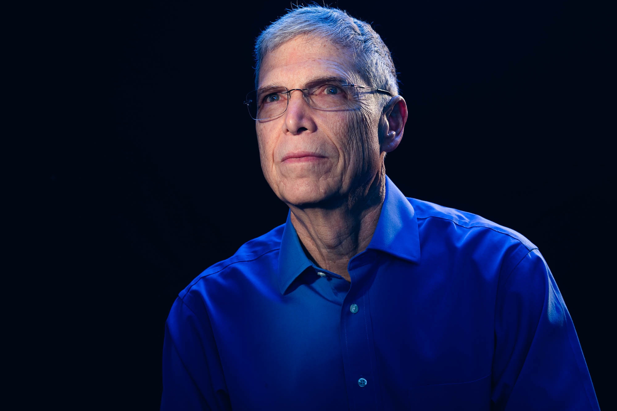 Gary Horowitz wears glasses and a blue shirt in a dark room