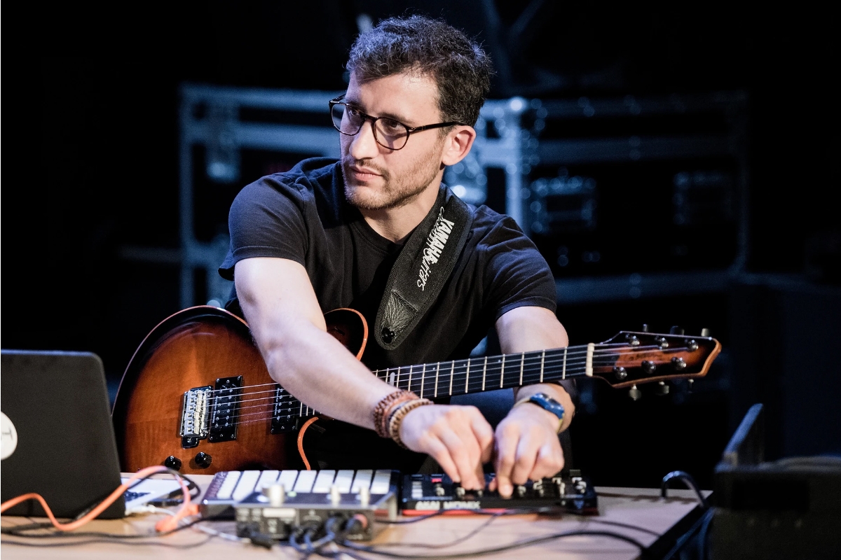 Diego Ratto turns knobs on a controller in front of a laptop with a guitar strapped to his shoulder