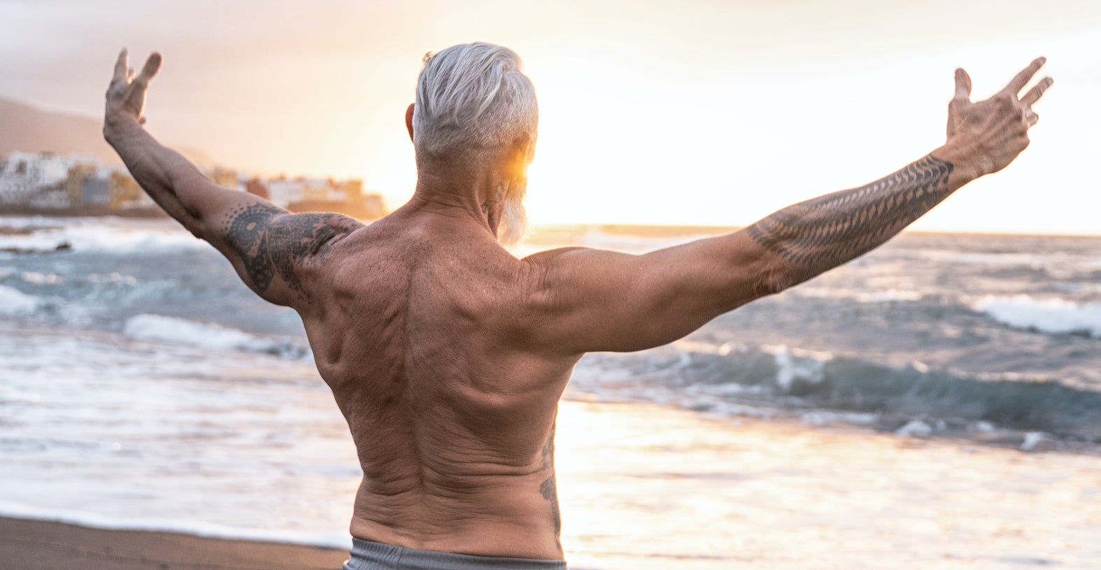 Man stands shirtless with his arms open towards the ocean