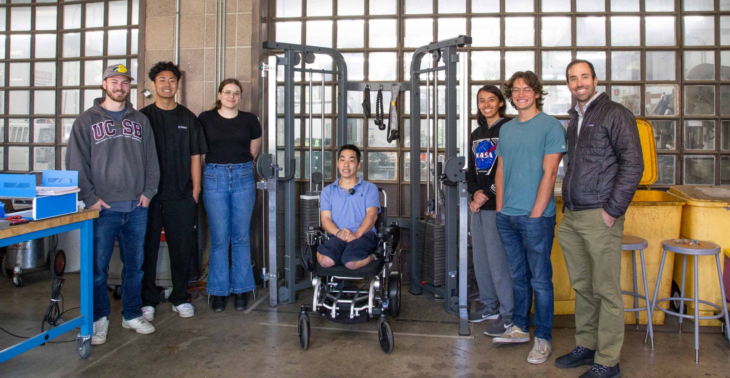 University students and professor flank an adult in a wheelchair in front of a weight machine