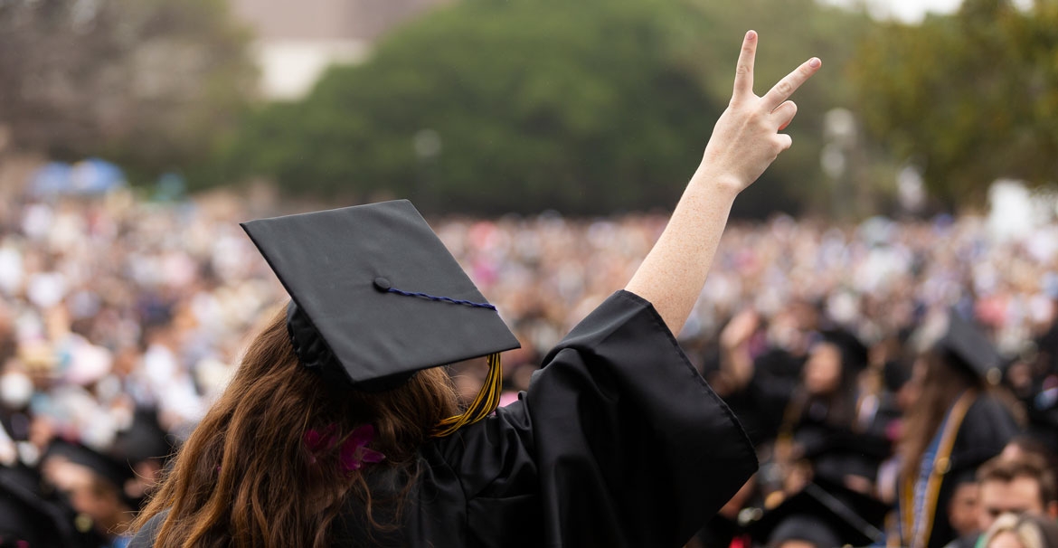 A graduate with long hair, wearing cap and gown, seen from back facing crowd of graduates