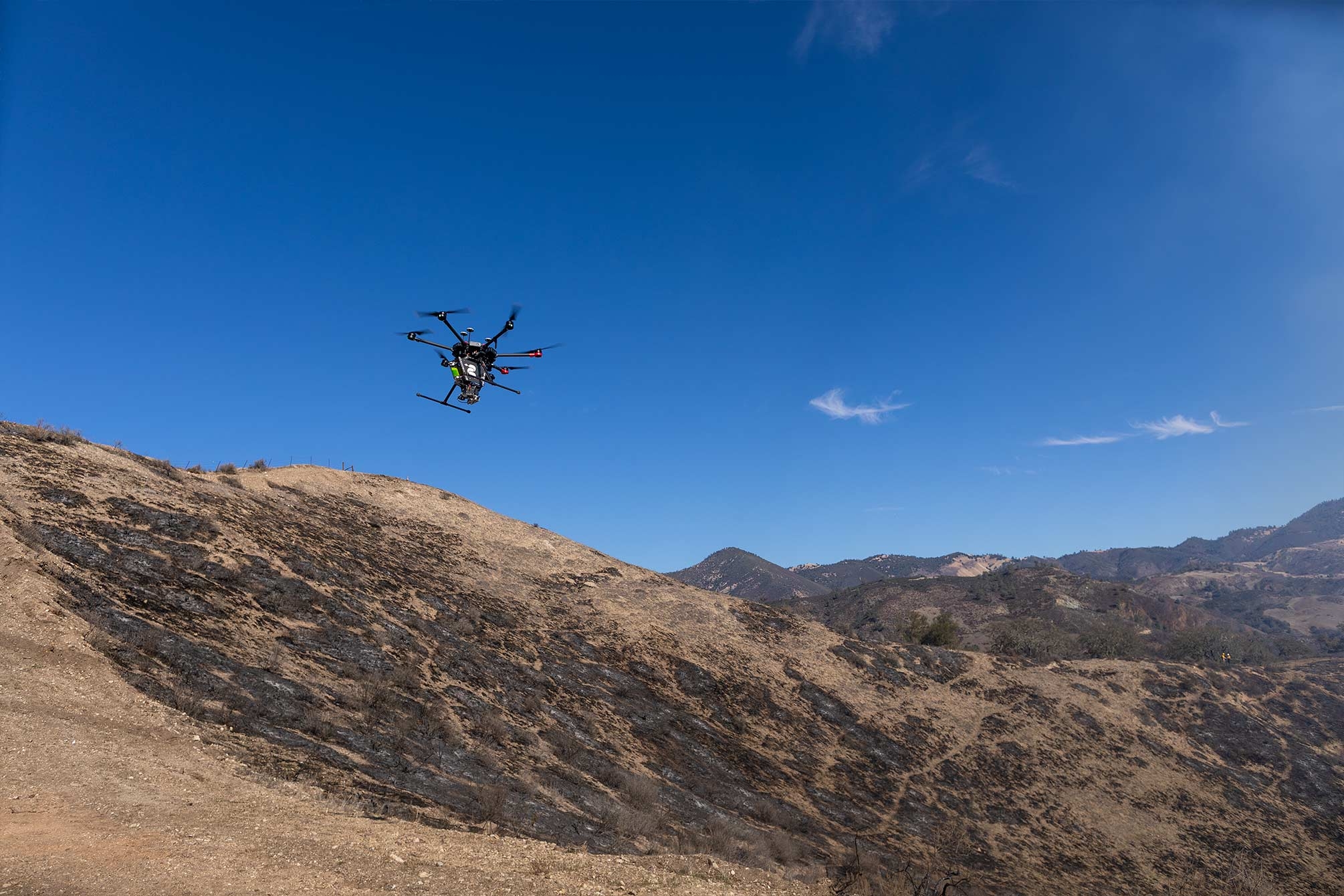 A hexacopter takes off into the blue sky above a scorched hillside.