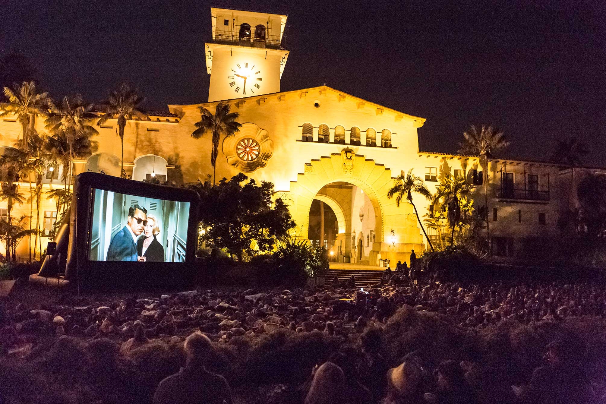 a courthouse at night with many people watching a film from the lawn on a large outdoor screen