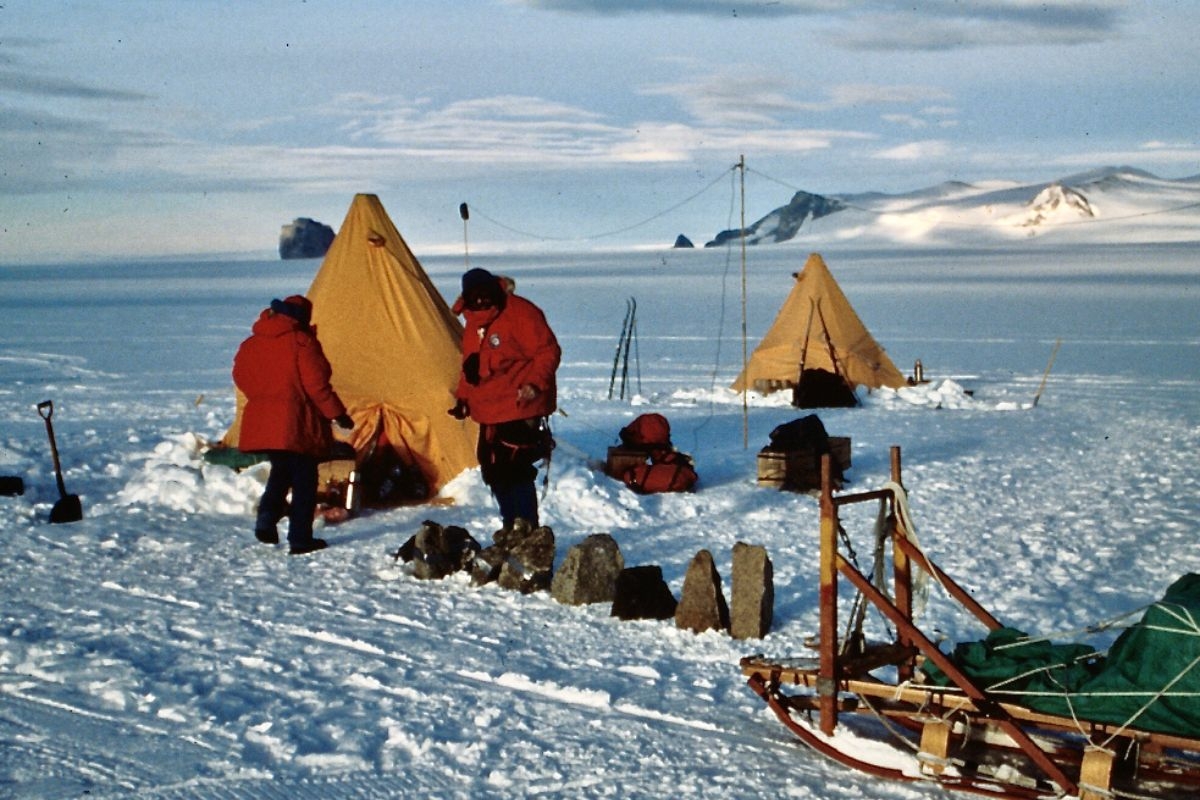 Two team members attend to camp surrounded by supplies, rock samples, tents and a sledge, with mountains in the distance.