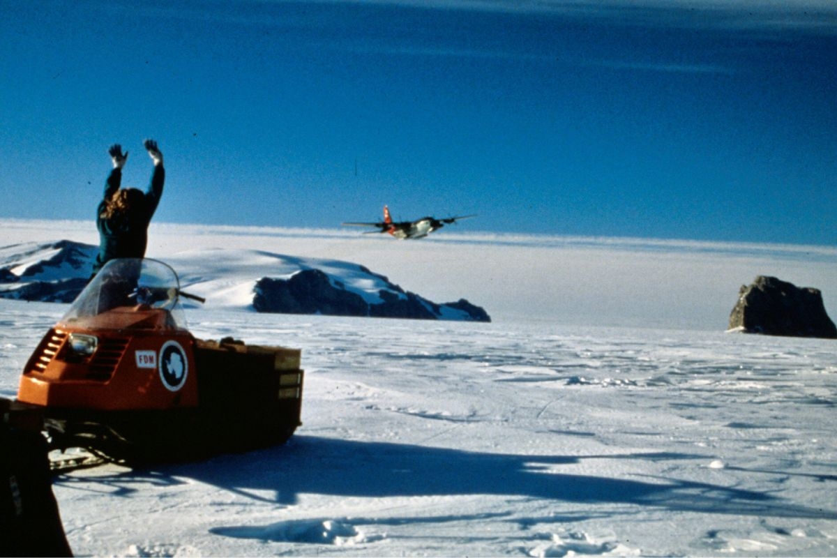 A researcher waves from behind a snowmobile as a cargo plane takes off in the distance.
