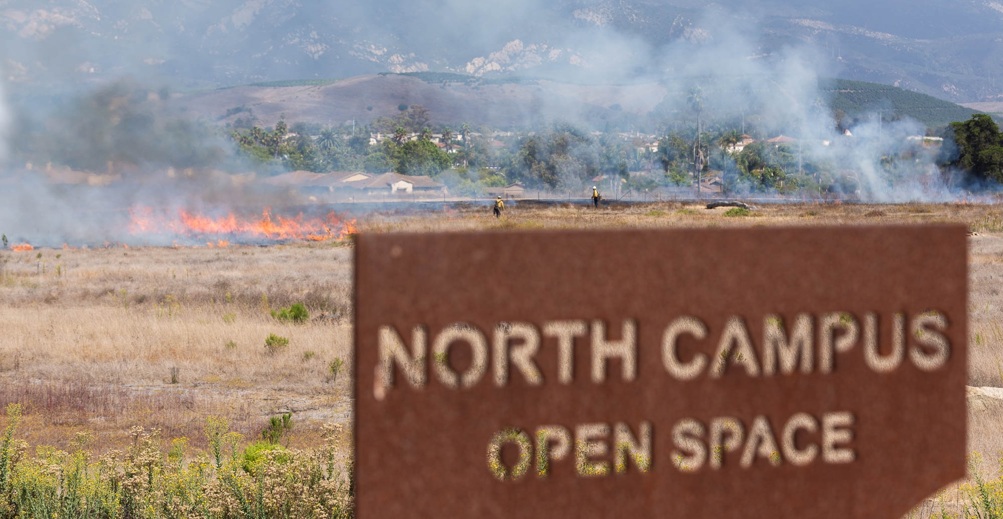 Flames burn behind a sign for the North Campus Open Space while firefighters manage the burn in the background.