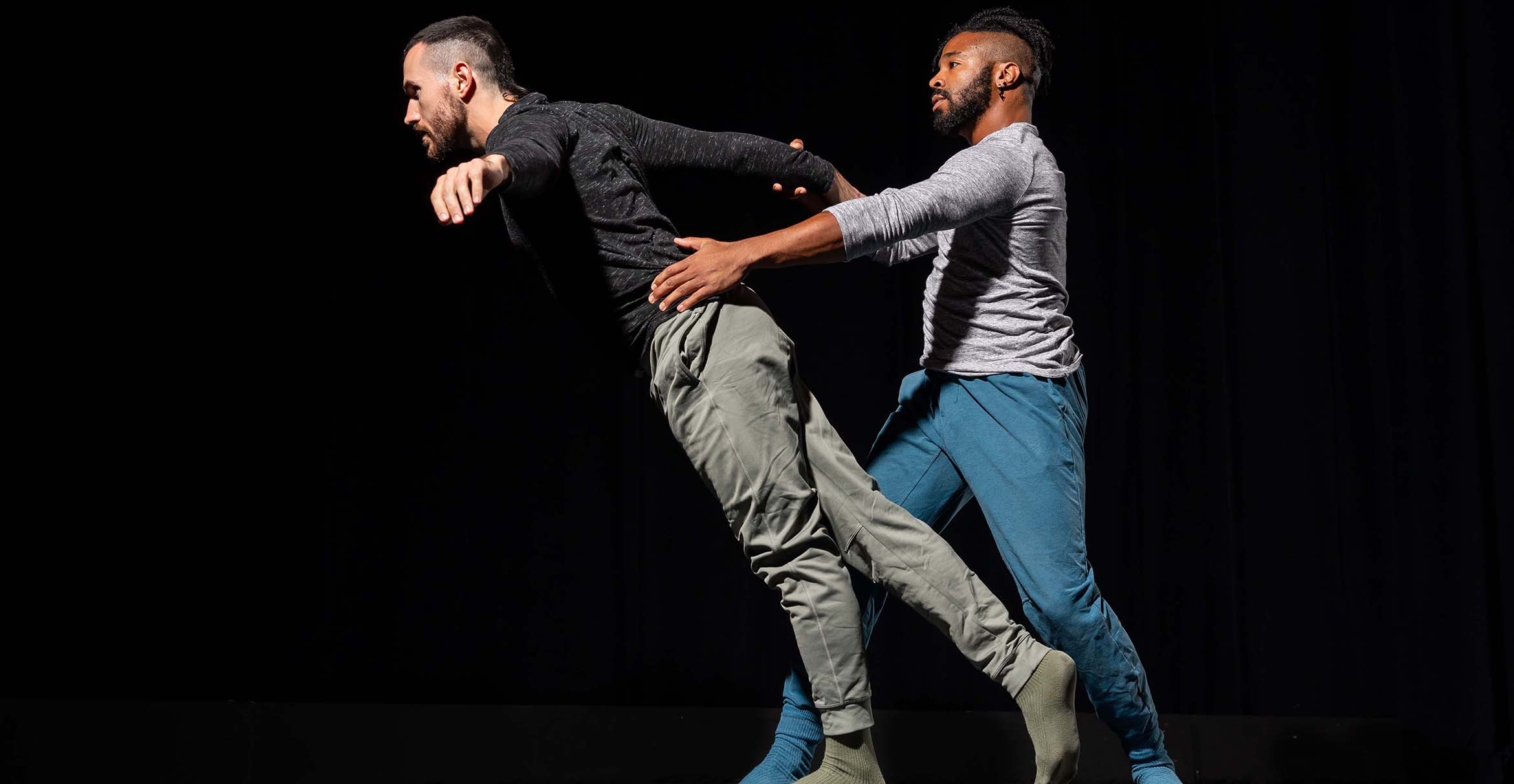 Two male dancers perform on a blackened stage