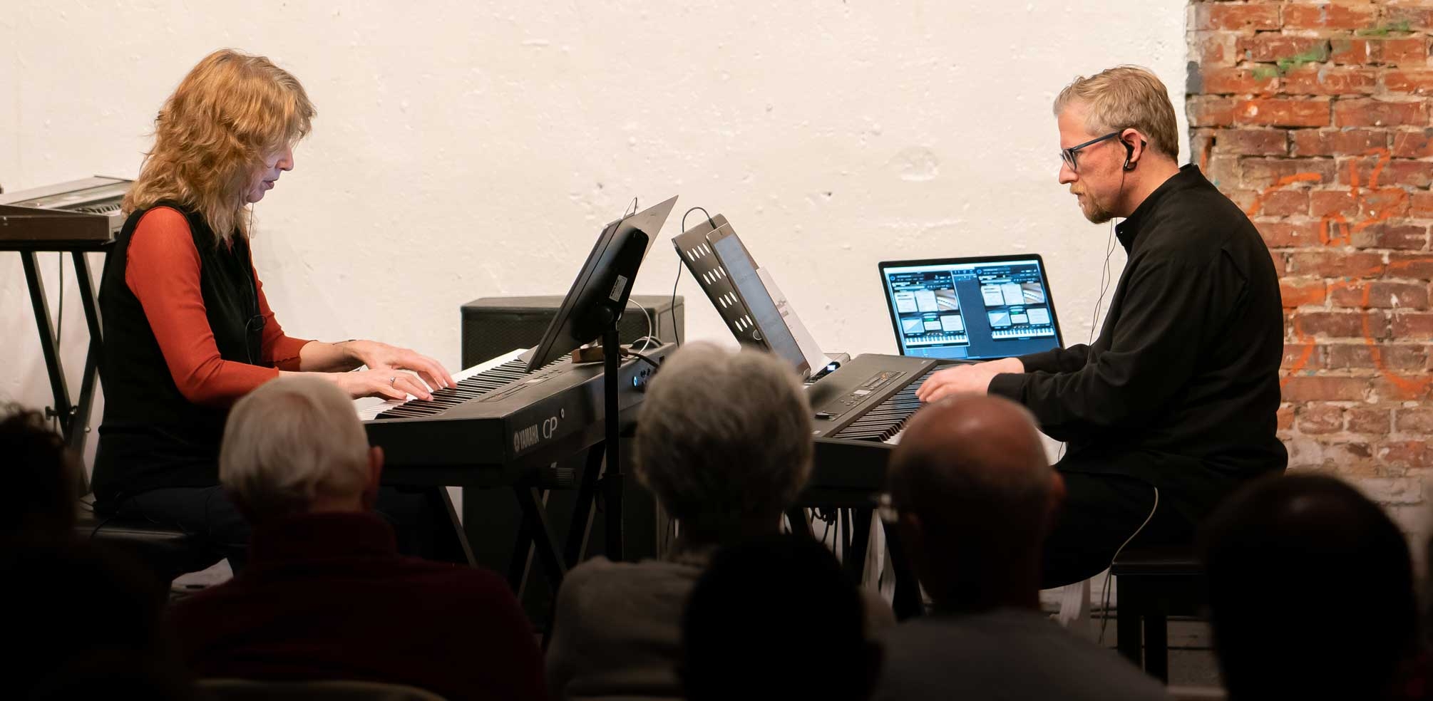 Two pianists play facing one another in a bright room, with an audience looking on