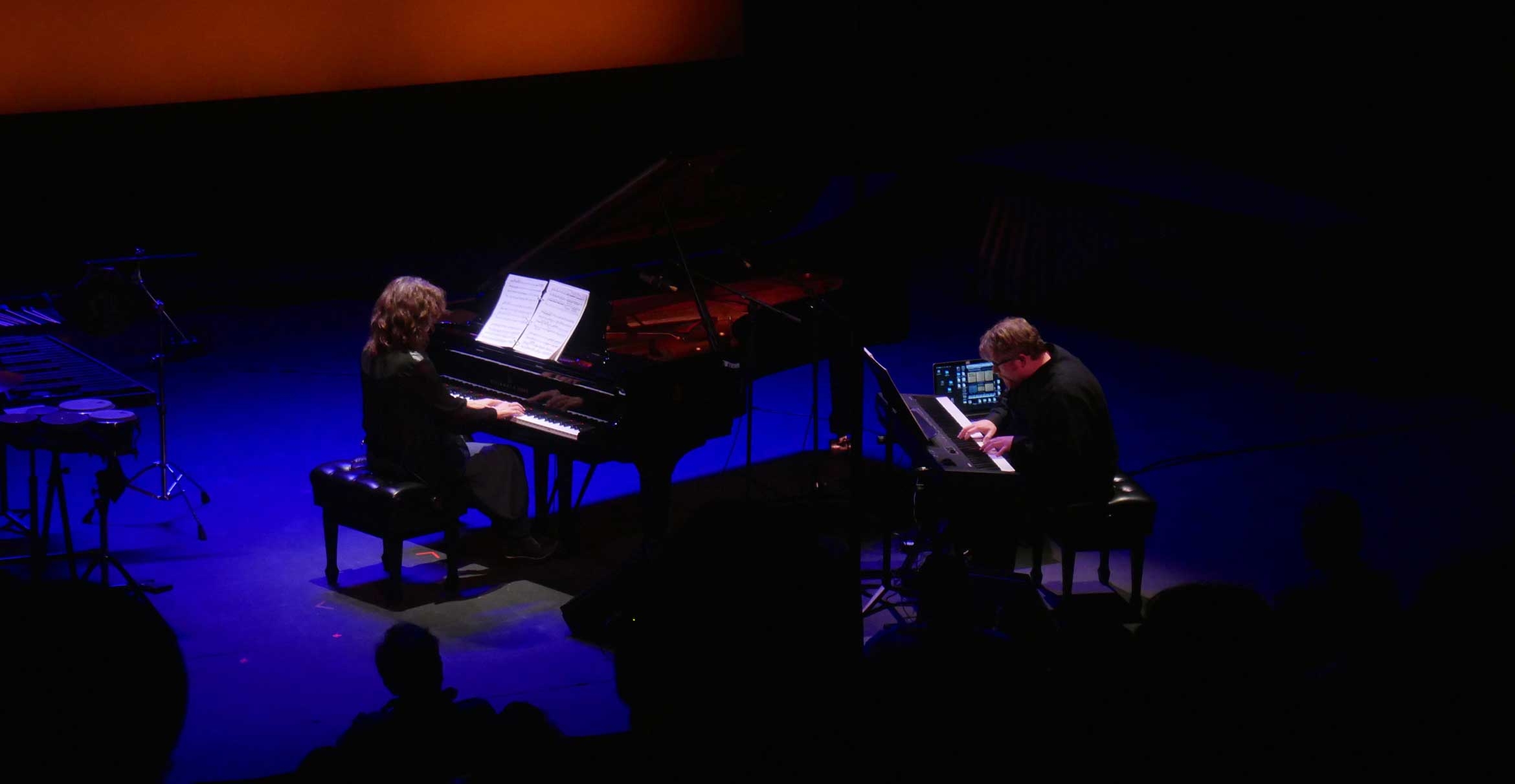 Two pianists play facing each other on a darkened stage