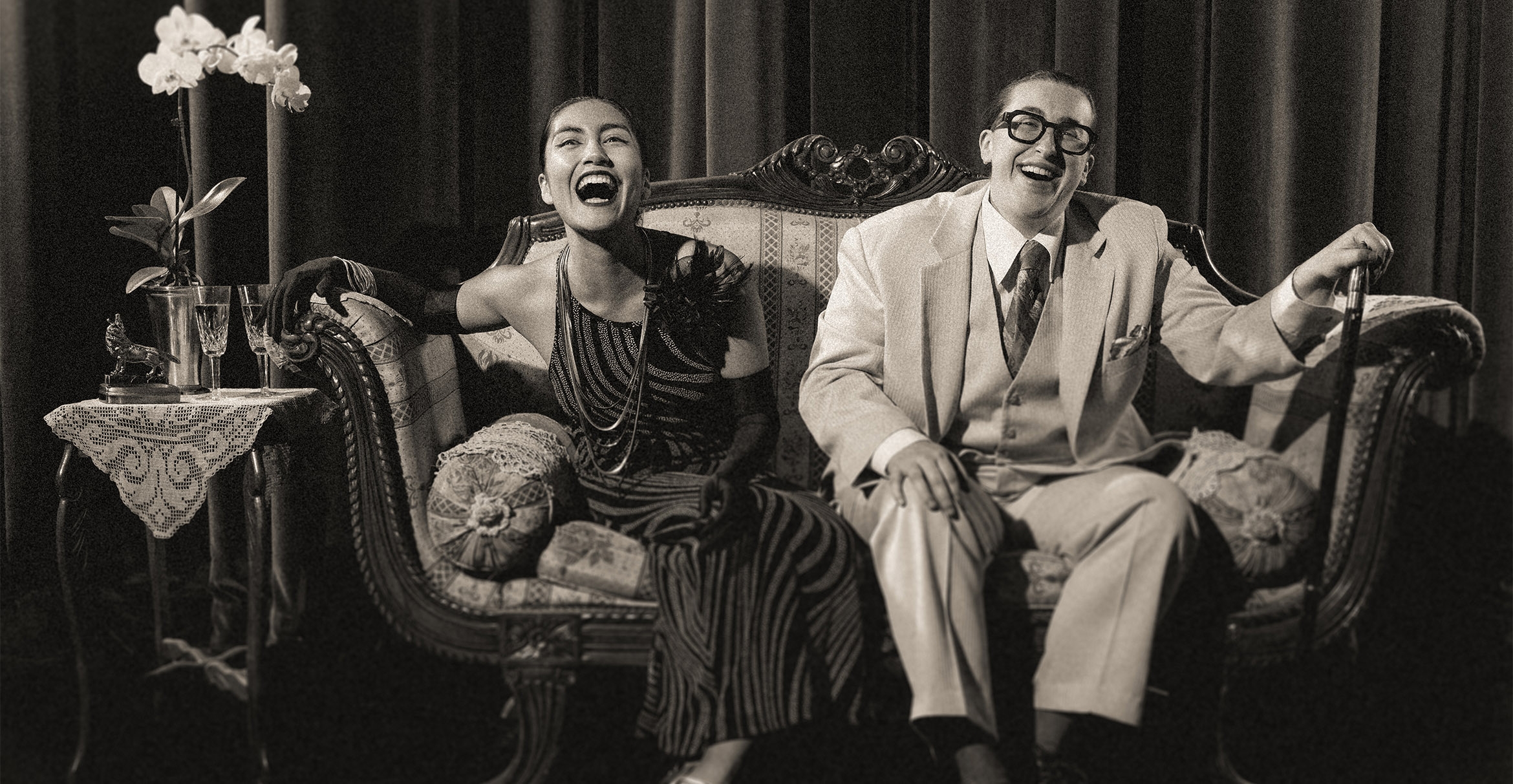 Two actors in 1920s attire sitting on a couch, laughing, in a black and white image