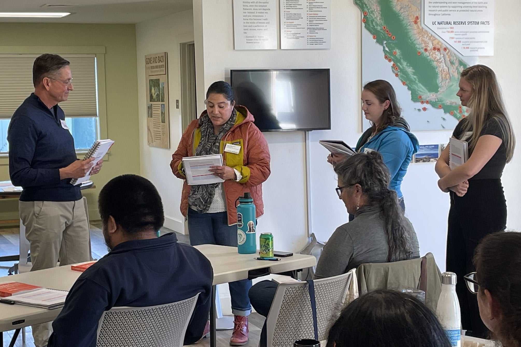 A diverse group of adults in a classroom, some seated and some standing, hold workbooks