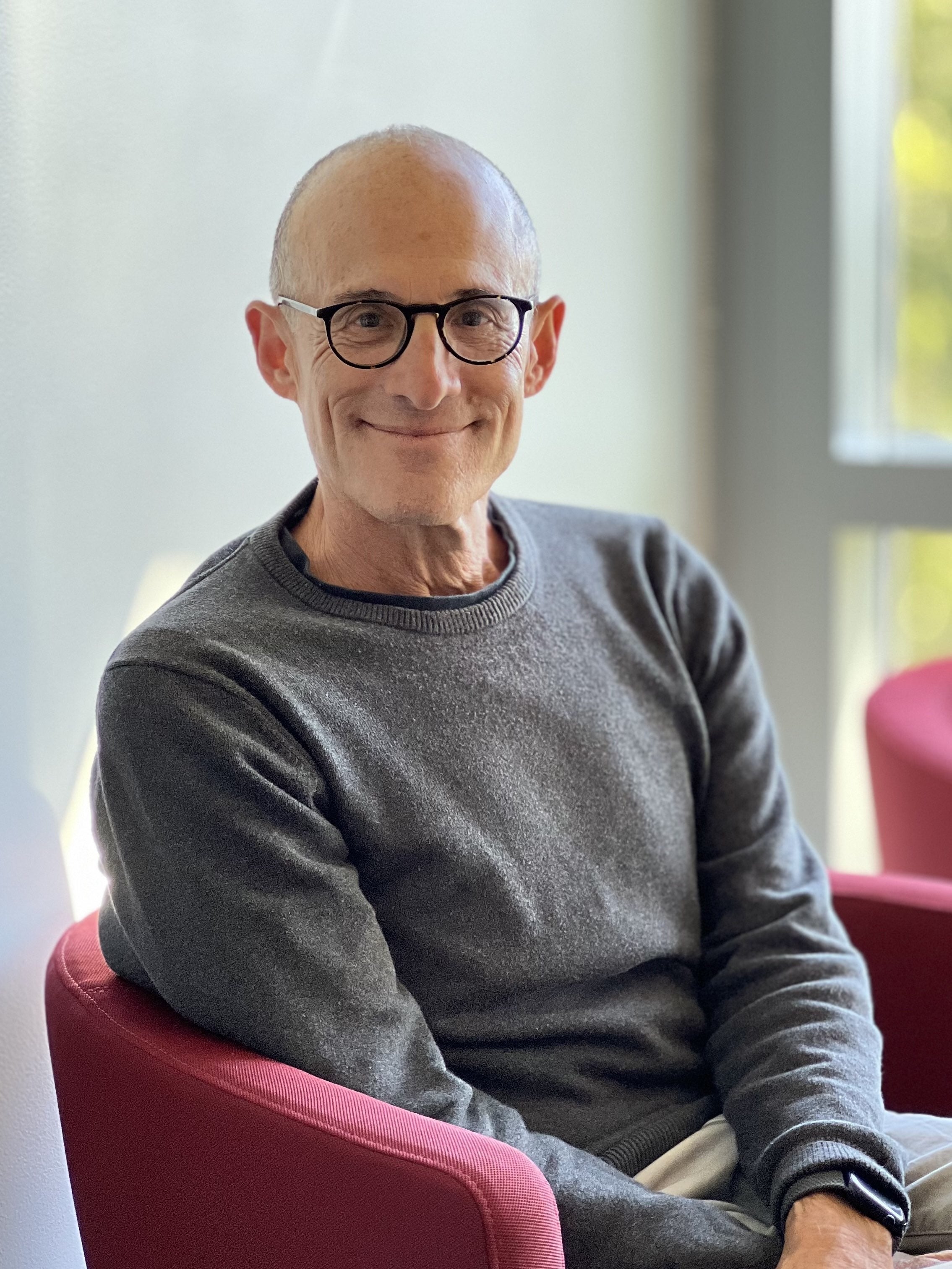 a man in a gray sweater with eyeglasses seated in a red chair