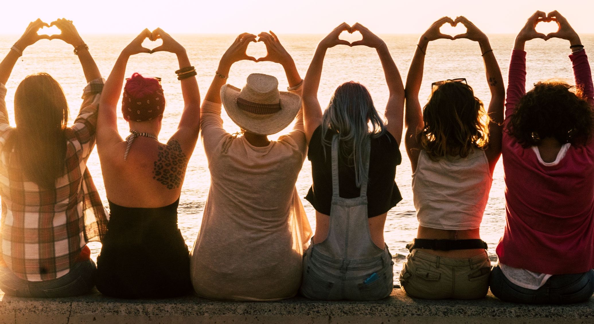 six women facing the sea with their arms raised in heart hands