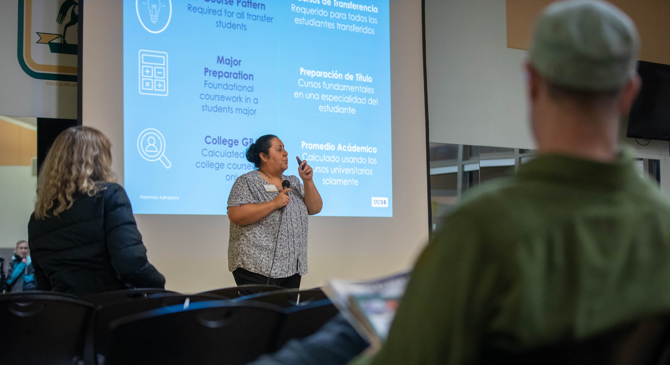 A woman standing in front of a large projection screen, holding a microphone and speaking to an audience