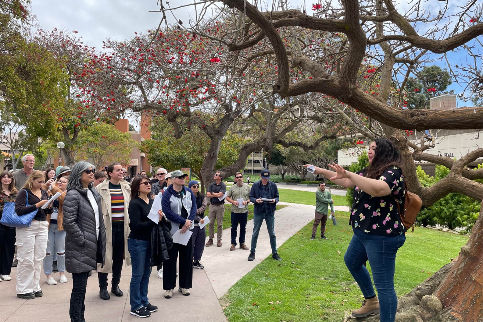 A diverse group of adults listening to a presentation outdoors, surrounded by trees