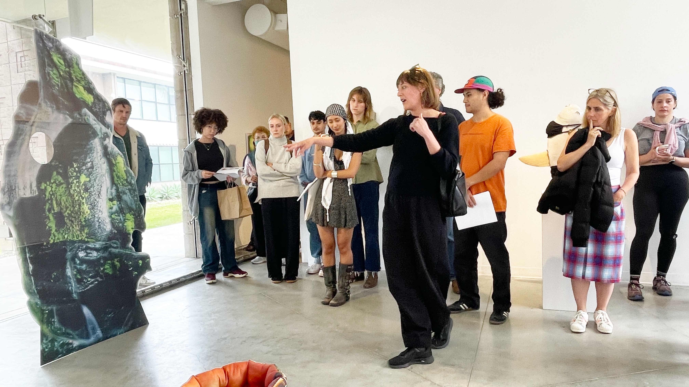 White woman points at a cardboard cut out in in a gallery with about a dozen people listening