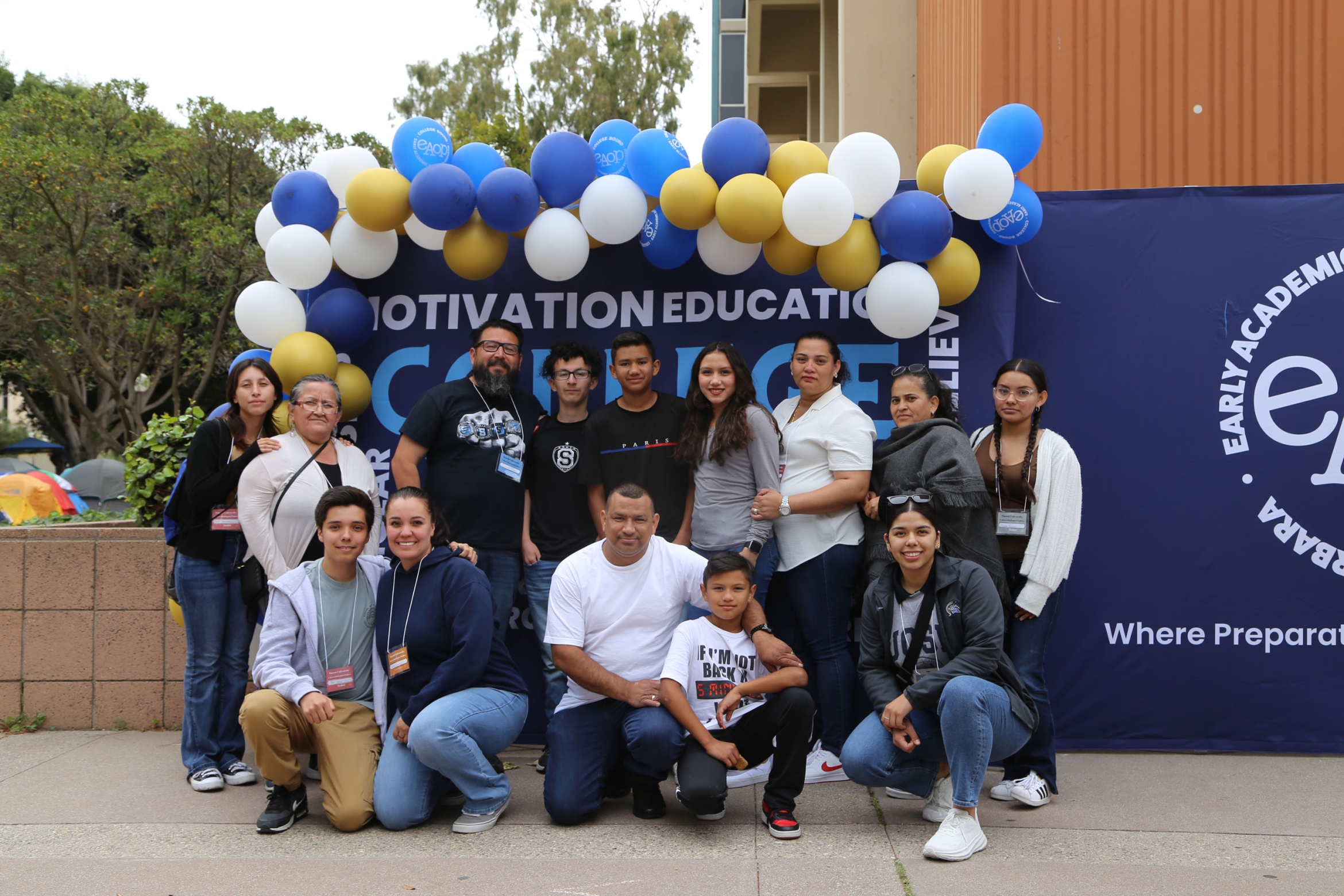 A diverse group of adults and children pose in front of a banner and balloons