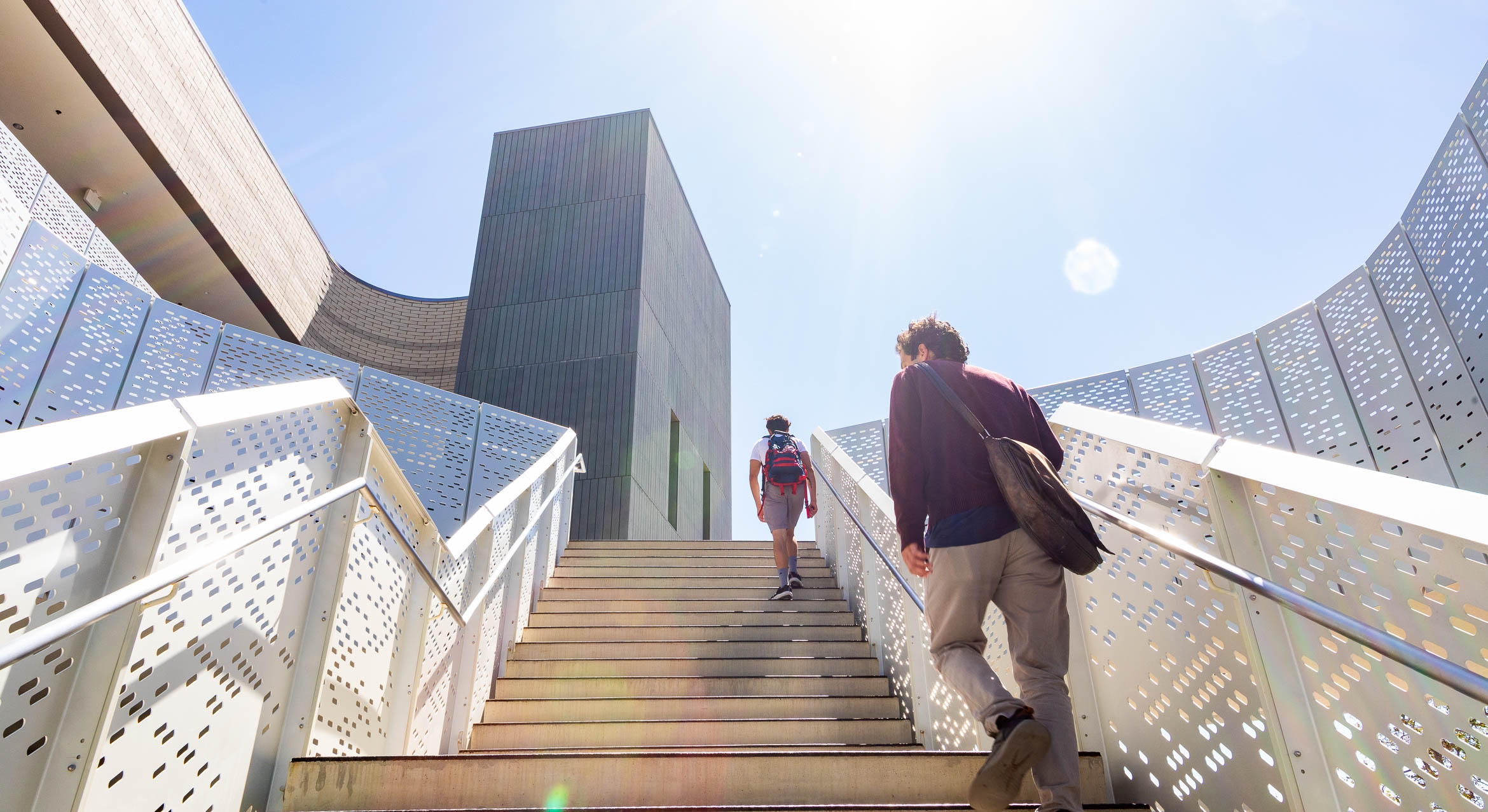 Two students ascend the modern stairway of an academic building with bright sun overhead