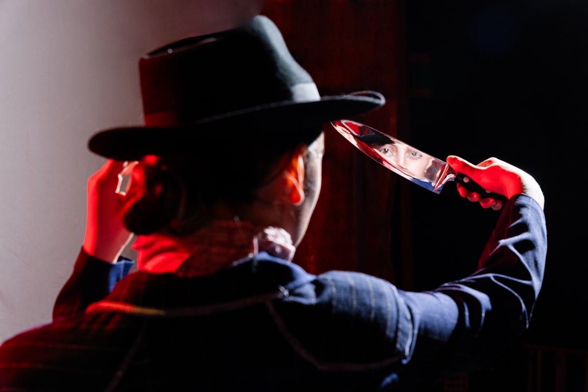 An actor on a darkened stage seen from behind, looking at their own reflection in a shiny knife