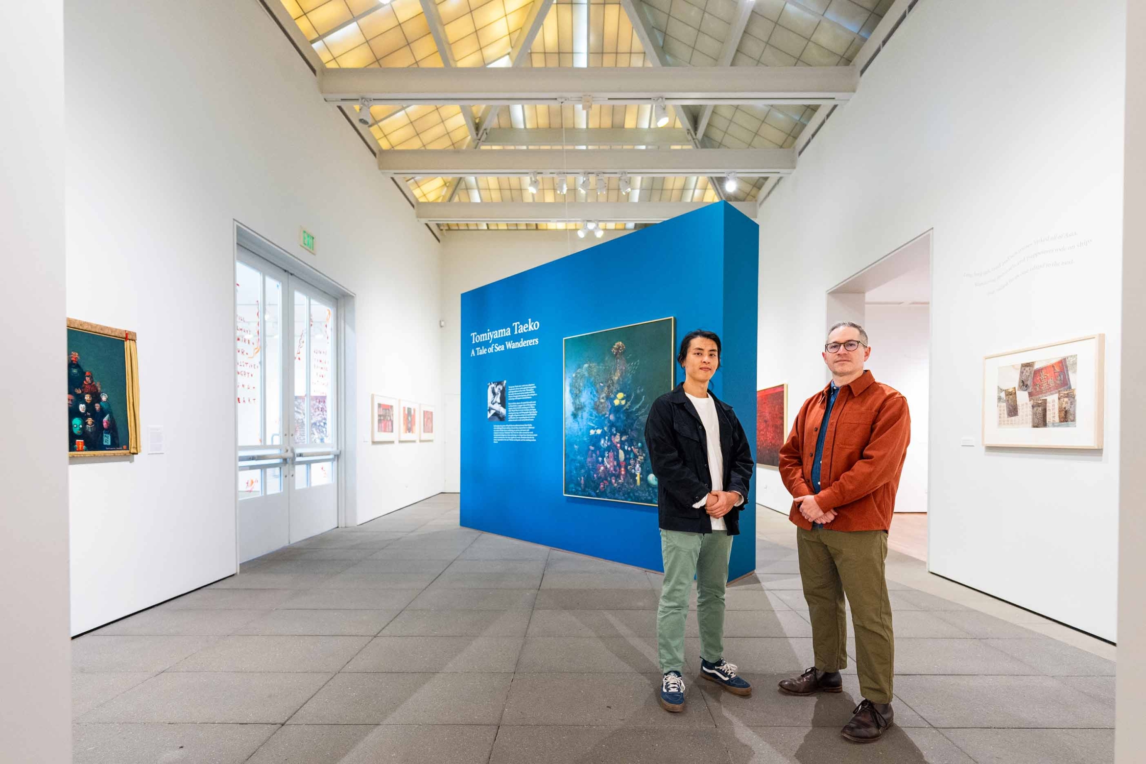 Two men inside the exhibition space with a blue floating wall behind with introduction text for the exhibition of Tomiyama Taeko