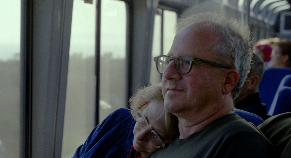 A man with glasses sits on a train, gazing out the window with a contemplative expression. A woman leans her head on his shoulder, partially obscured, wearing glasses and a blue sweater. The train interior is softly lit, and other passengers can be seen in the blurred background.