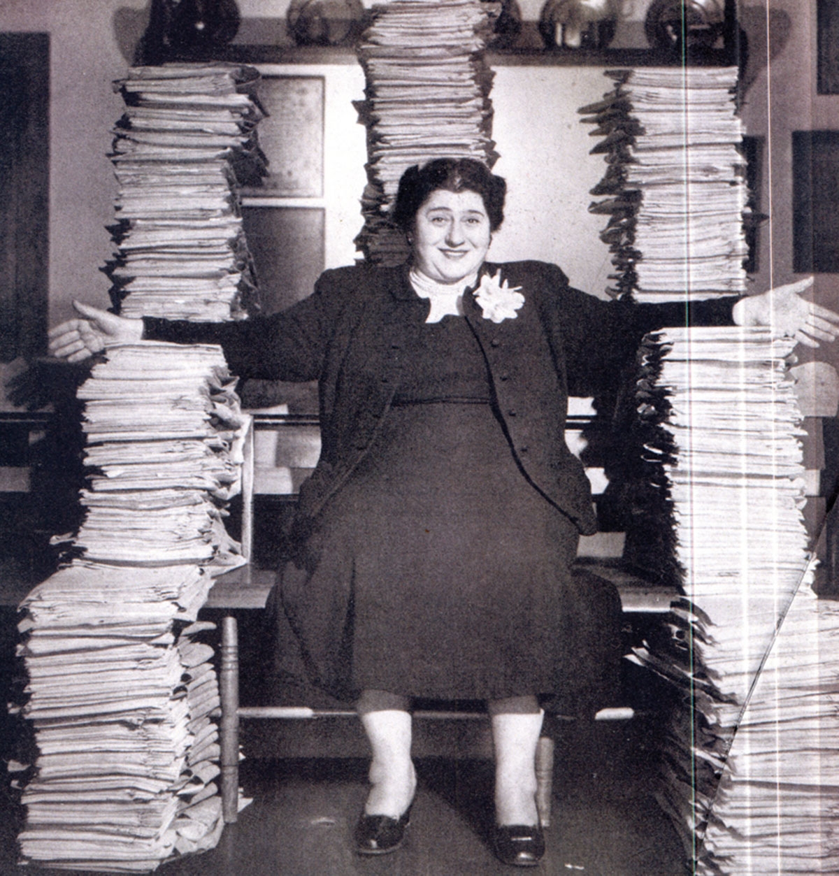 Black and white photo, circa 1950s, of a woman in a dark suit, seated, surrounded by scripts