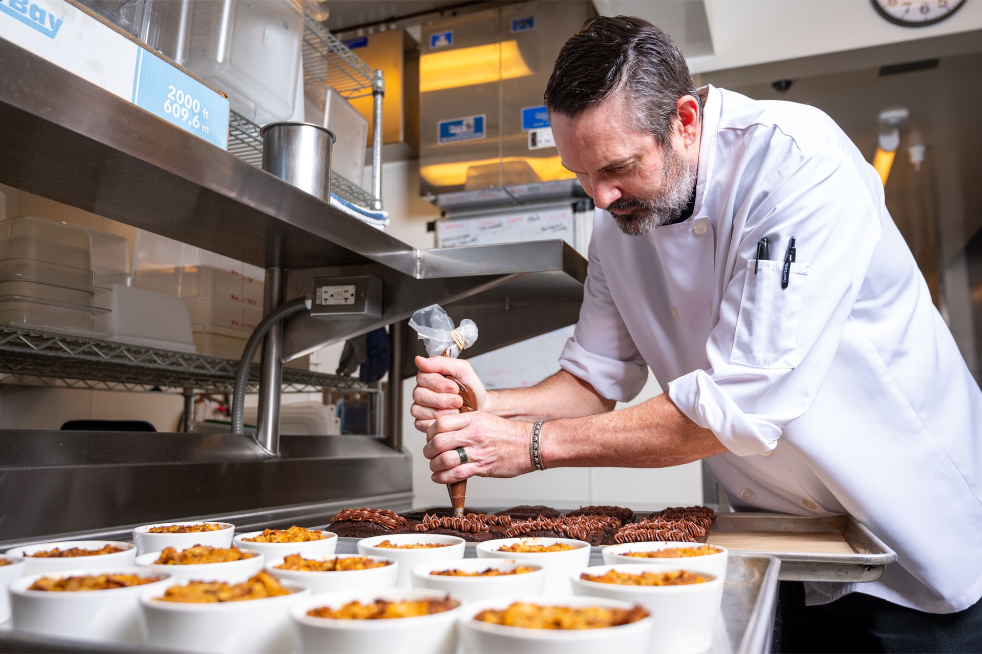 A male chef in a white chef's coat leans over trays of food holding a piping bag, frosting cakes