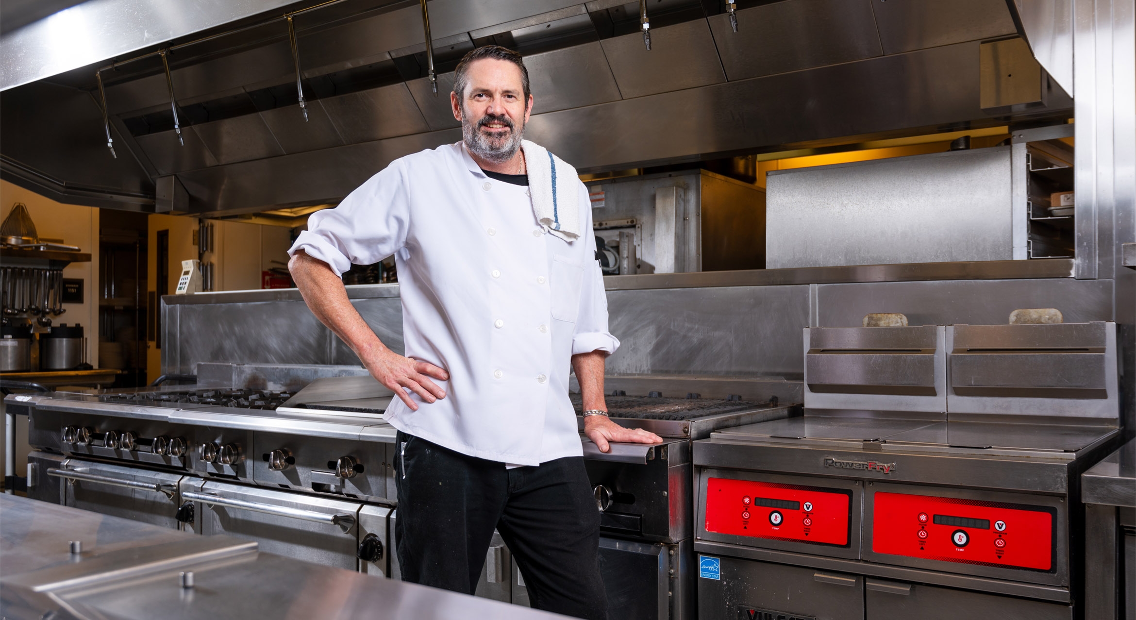 Portrait of a male chef, with dark hair and a greying goatee, in an industrial kitchen