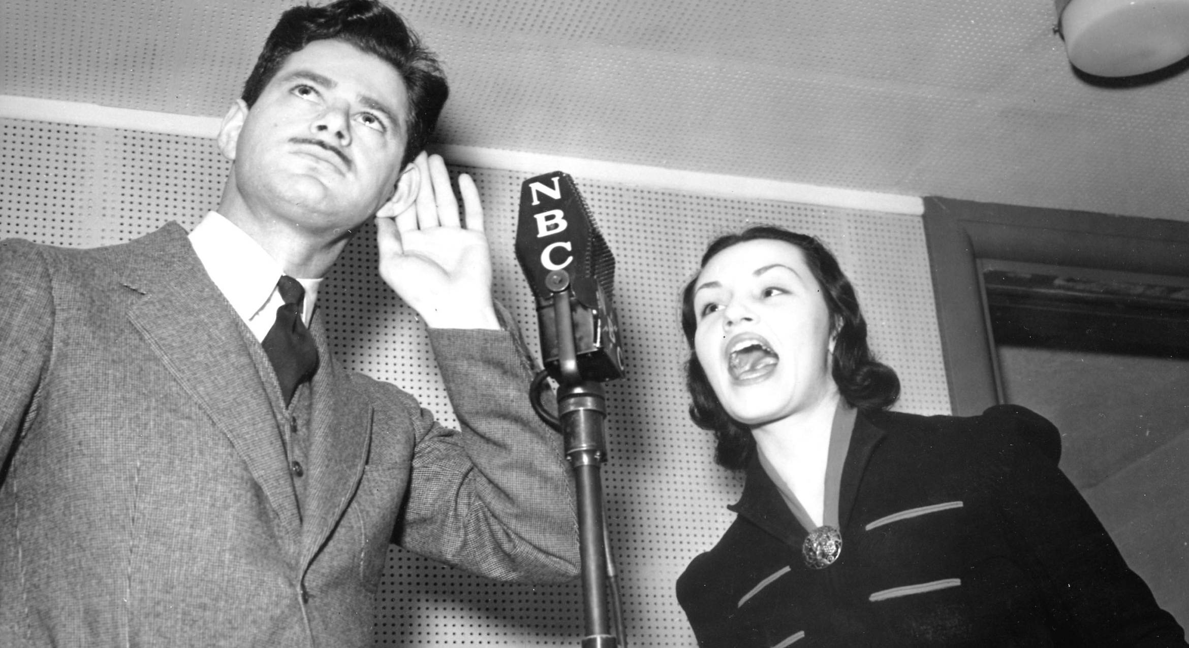 A man and a woman behind a standing microphone, circa 1930