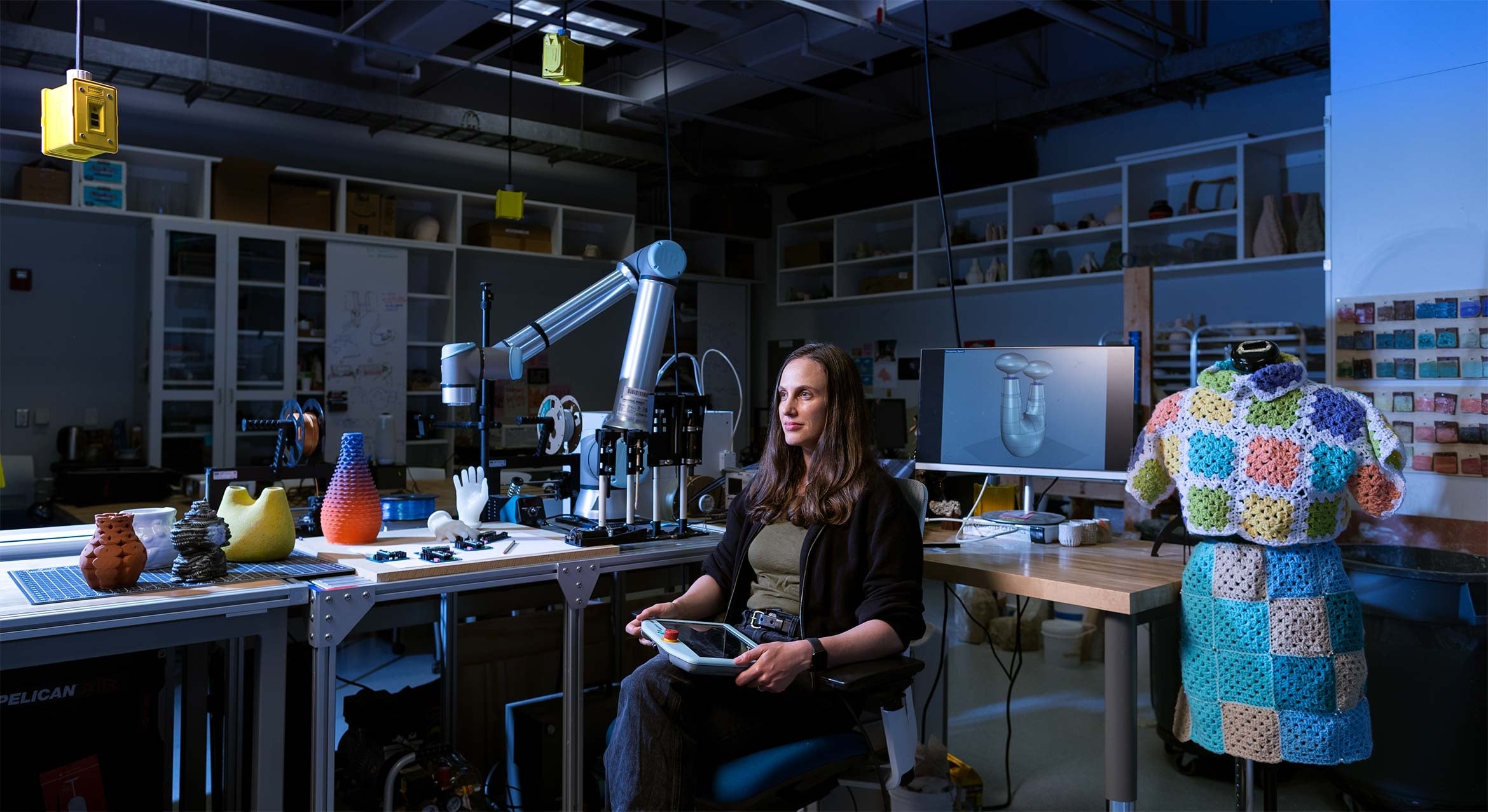white woman sits at desk with tablet, on desk is robotic arm, coiled clay vessels and computer screen, crochet dress on mannequin at side