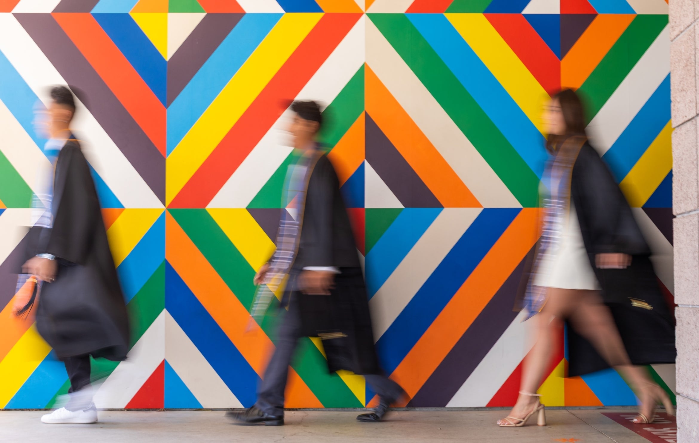 Three people in graduation gowns walk past a colorful background of geometric shapes