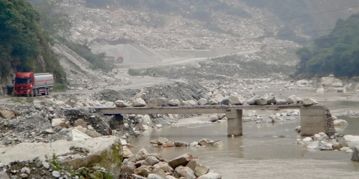 Boulders are strewn across the remains of a bridge in a river valley. Debris is all around, as trucks navigate a cleared roadway.