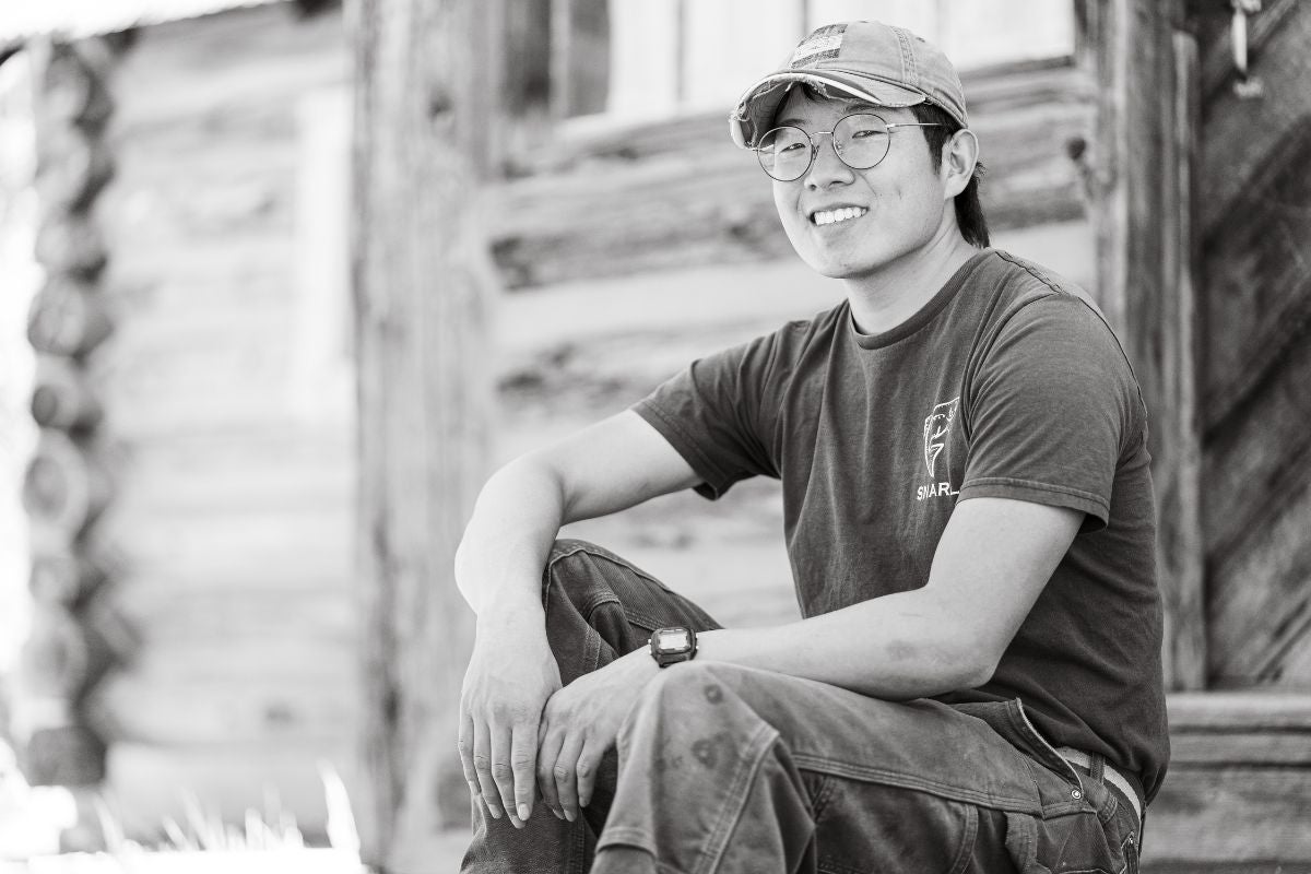 Juyung Yoo sits on the cabin steps in a reserve t-shirt, with large, round spectacles beneath his tattered cap.