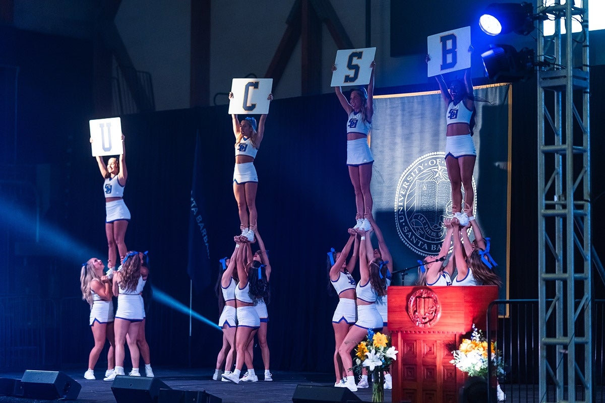 Four clusters of cheerleaders in white uniforms hold up signs reads UCSB