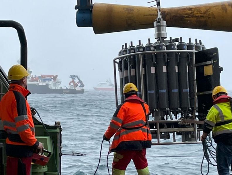 Three researchers in high visibility gear deploy a large piece of equipment from the deck of a vessel, with two other research vessels in the foggy background.