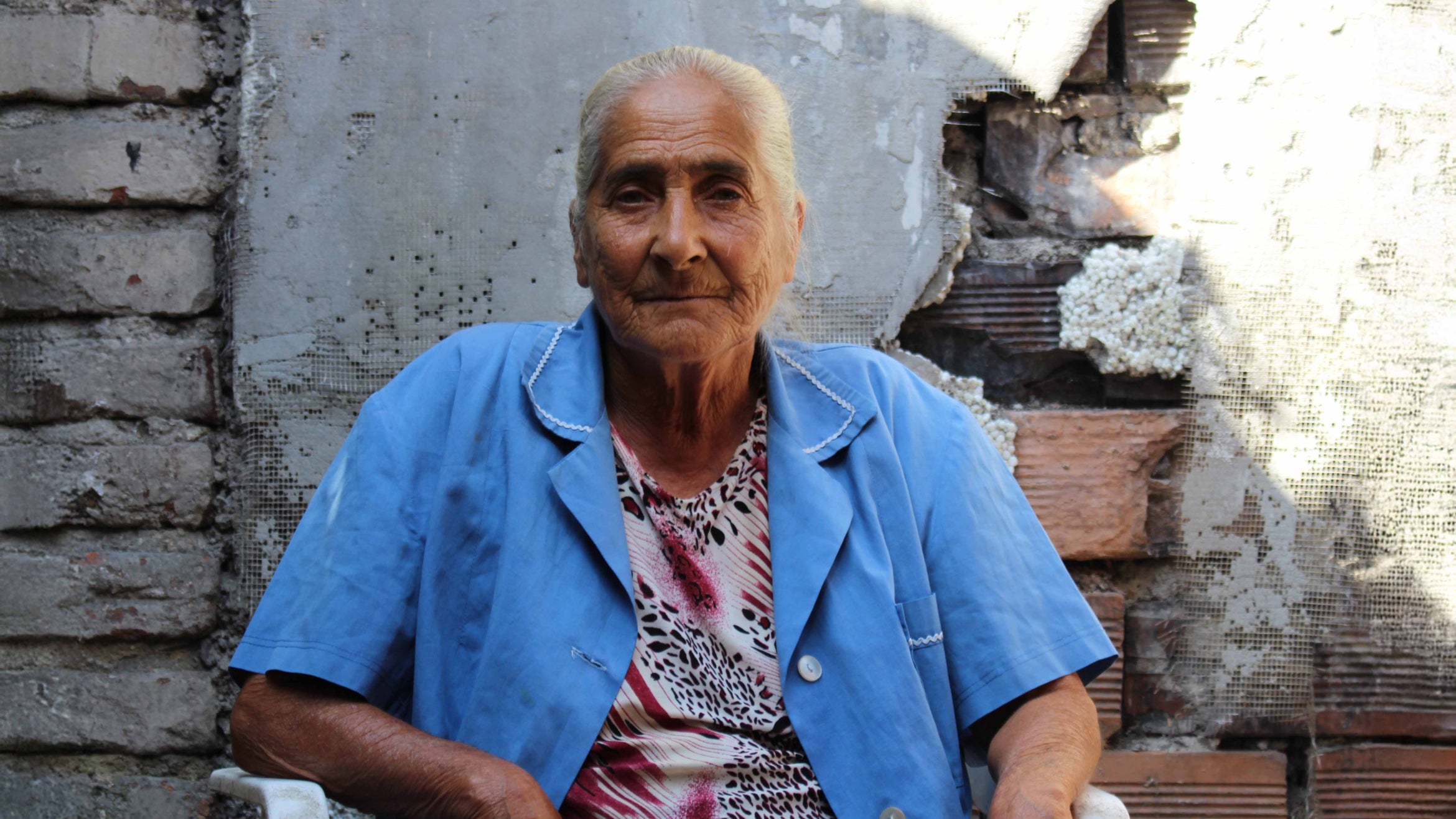 Older woman with grey hair and blue coat sits outside rustic building