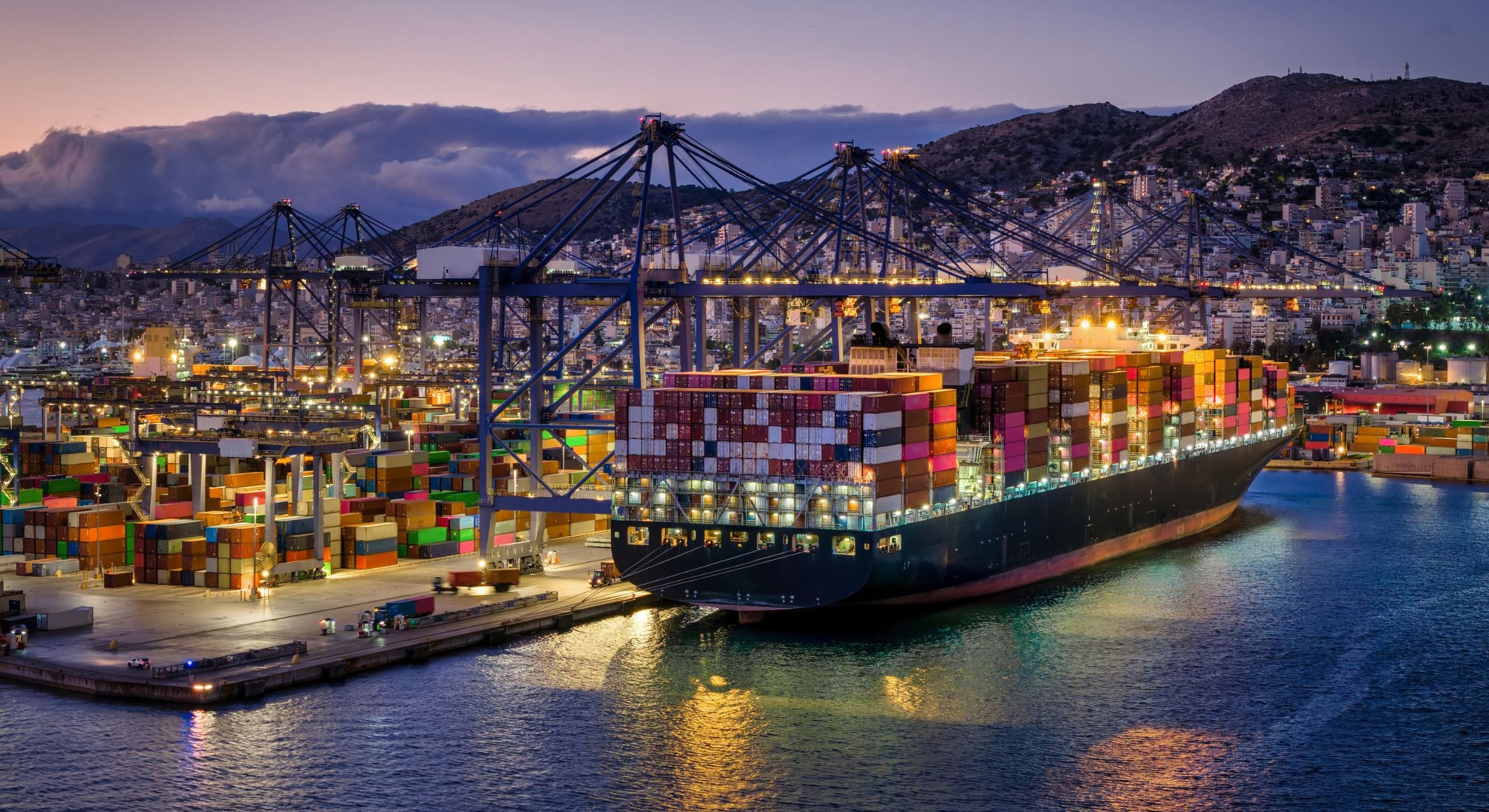 A super cargo ship is tended to by cranes and trucks in a commercial terminal. Lights from the port brighten the dimming sky, with mountains and low clouds in the distance.