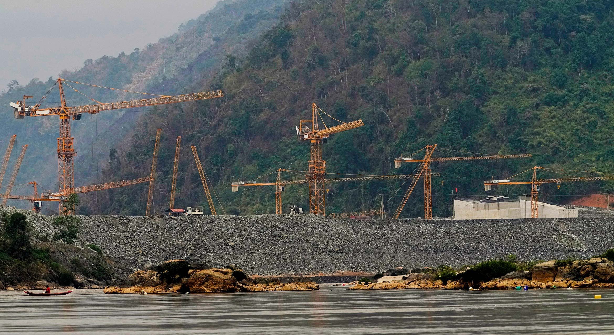 A fisherman catches fish in front of an earthen embankment with a dozen yellow construction cranes peaking up behind it, with lush forested hills in the background.