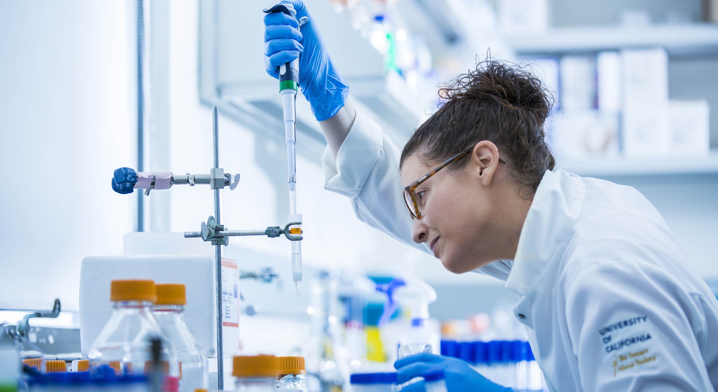 Woman wearing a lab coat, glasses and blue gloves drips liquid into a test tube