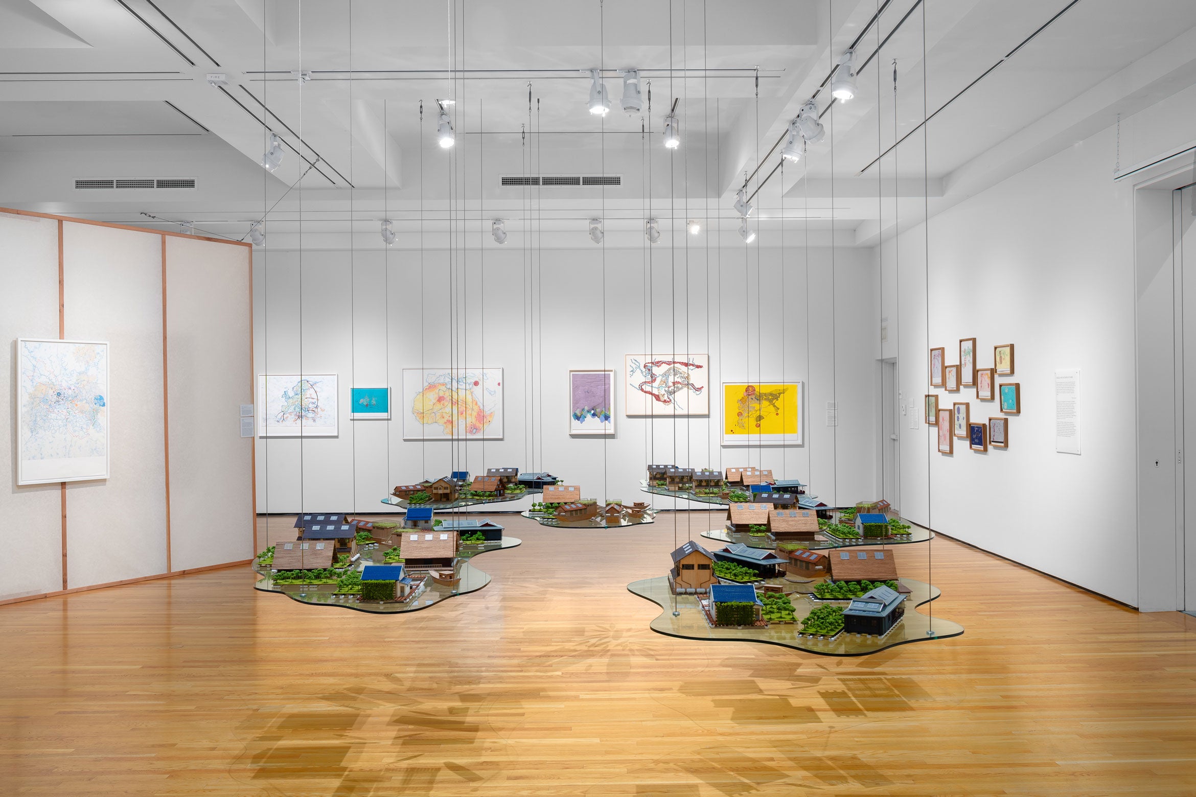 inside an exhibition in a museum with hanging maquet of homes or neighborhood with biombo and white walls surrounding with artworks on the walls