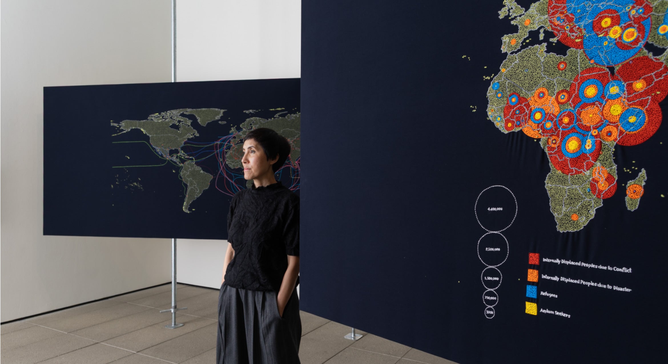 woman with short black hair in black and grey clothes stands near big black rectangular maps with brightly colored embroidery maps