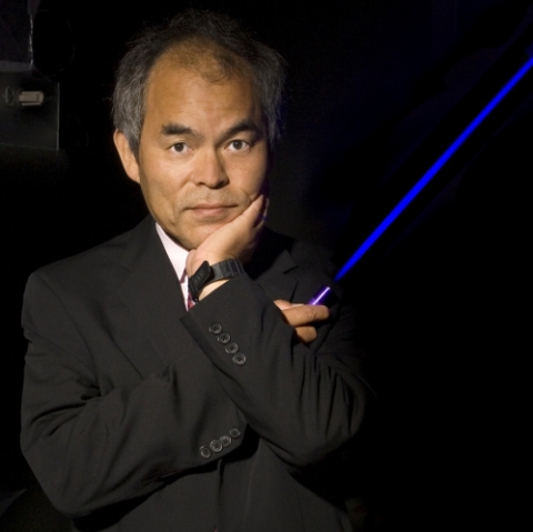 Shuji Nakamura, wearing a black suit, holds a laser in one hand and rests his chin on the other hand