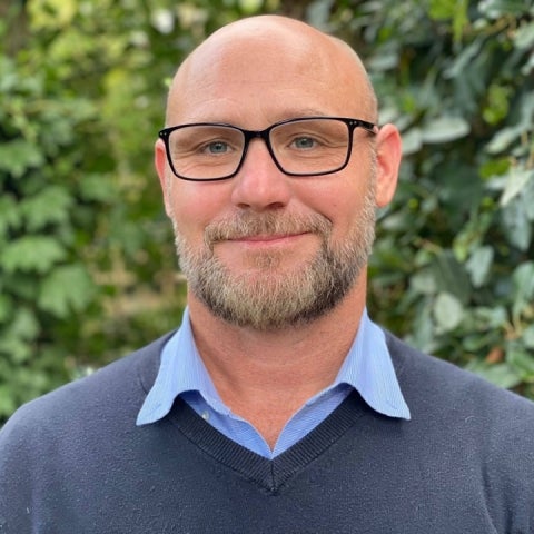 Jeffrey Hoelle stands outside wearing black glasses and a sweater over a blue collared shirt