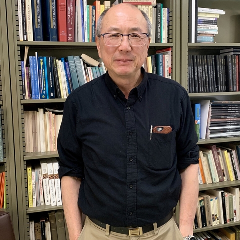 Alan Liu stands in front of a bookshelf wearing a black collared shirt and glasses